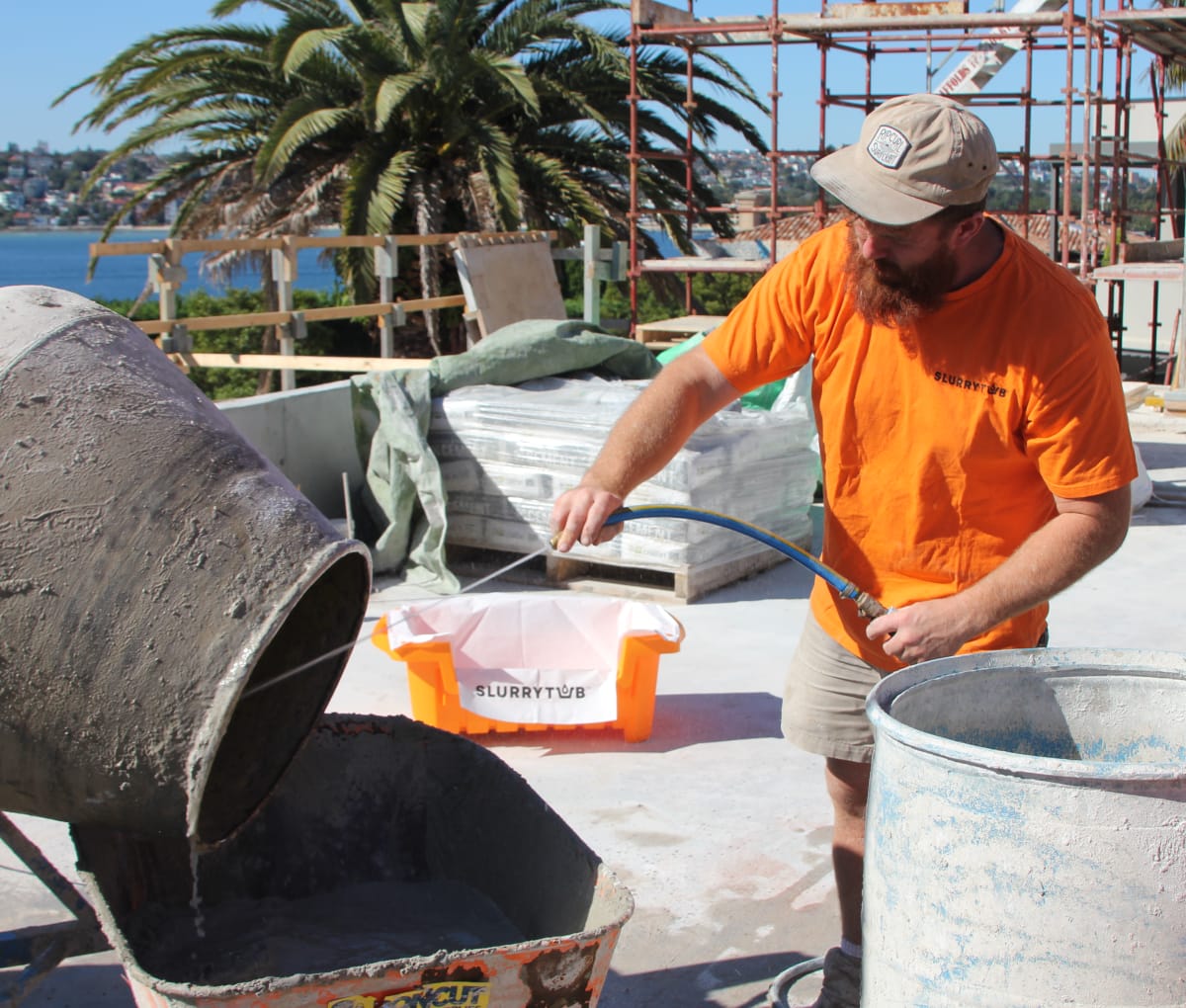 Man in hat on a sunny construction site with palm trees in the background rinsing a concrete mixer into a slurrytub. 